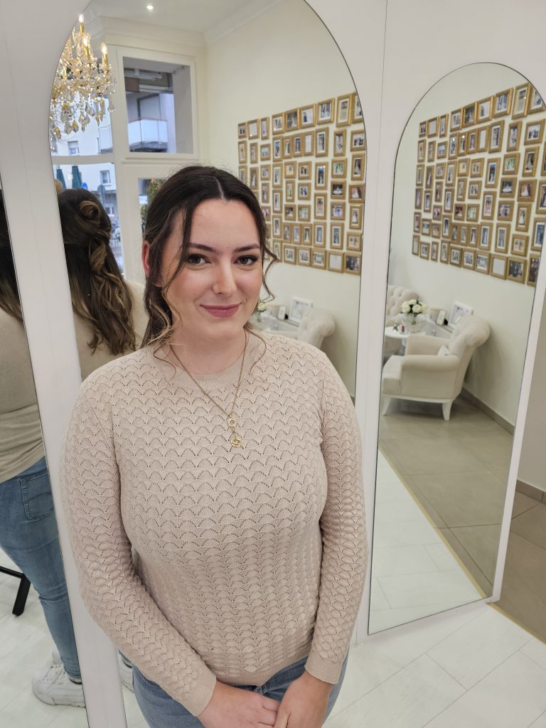 Eine junge Frau nach dem Braut-Styling durch Shiva Beauty Lounge in einem Brautmodengeschäft vor drei hohen Spiegeln.